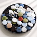 A top-down view of a round black tray filled with smooth, polished stone-like discs in assorted colors including white, blue, green, black, brown, red, and beige, set against a light neutral background with soft shadows.