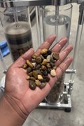 A hand holding a handful of polished river stones and gravel, with clear cylindrical columns containing water and sand filters in the background.